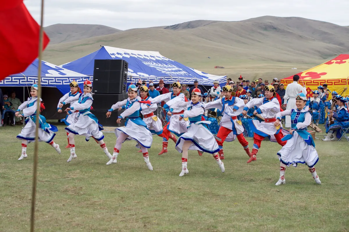 Dancers in white, blue and red costume and boots perform on grass in front of a blue patterned tent, with an orchestra and green hills behind them