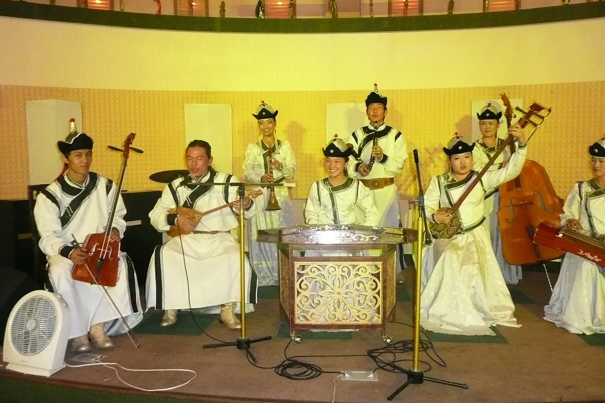 A Mongolian folk ensemble in white silk costume and black hats on a small stage, with a fiddle, a lute, a hammered dulcimer, a zither, wind pipes and a double bass