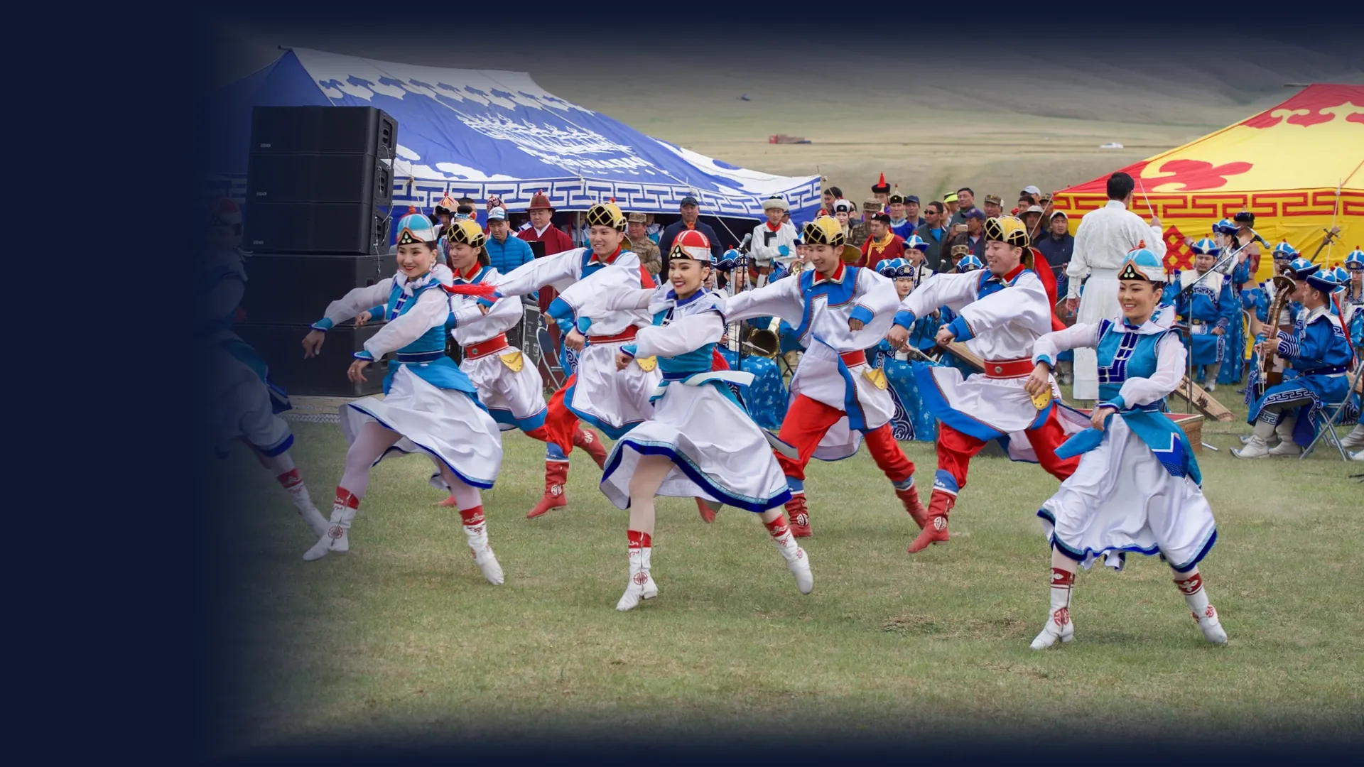 Dancers in white, blue and red costume and boots dance on grass in front of a blue patterned tent, with an orchestra and green hills behind them