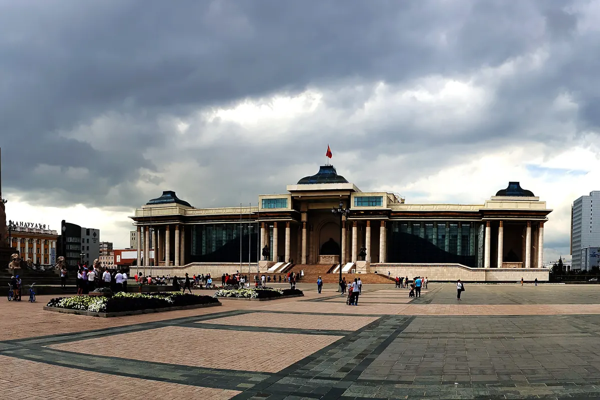 Sükhbaatar Square in Ulaanbaatar under a dark sky, with the columned Government Palace in the middle and a stone monument on the left
