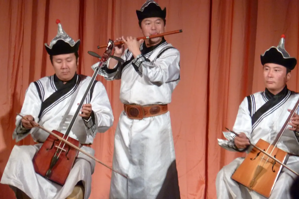 Three musicians in white silk robes and black felt hats play two horsehead fiddles and a flute in front of orange stage curtains