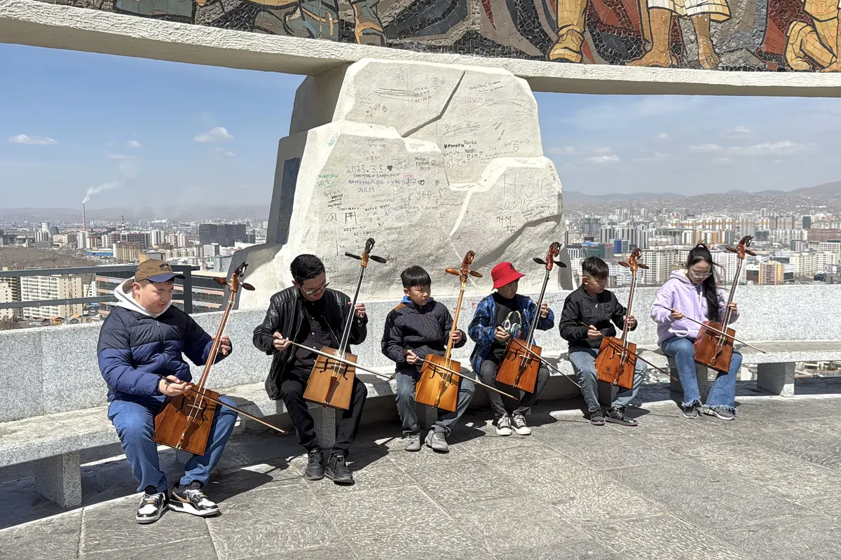 Six people of different ages sit on a granite bench playing horsehead fiddles at the Zaisan Memorial, with Ulaanbaatar apartment blocks and hills behind them